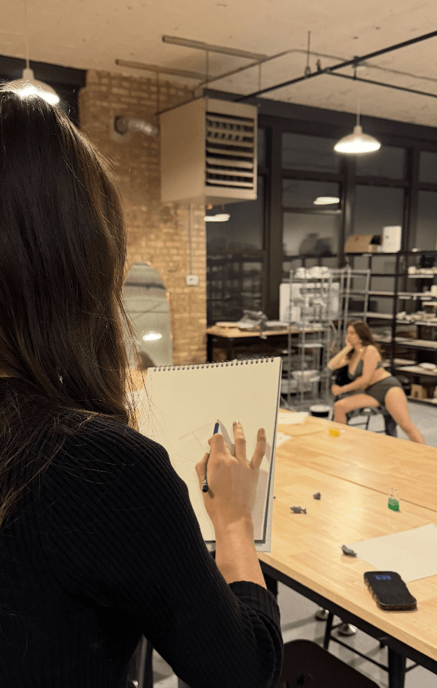 A student sketching on a pad while the live model poses in the background